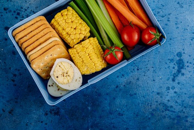 image of a lunchbox with crackers, boiled eggs, corn, veggies. on a blue background
